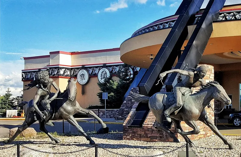 Photo: bronze sculpture of two riders on horseback in front of the curved entrance of Painted Hand Casino in Yorkton, under a blue summer sky.